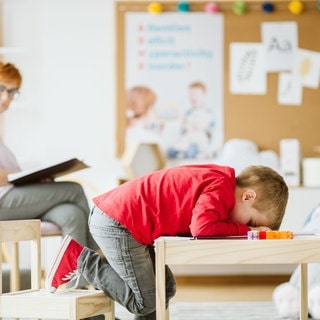 Kleiner Junge sitzt verzweifelt an einem Tisch. Frau im Hintergrund ist seine Therapeutin.