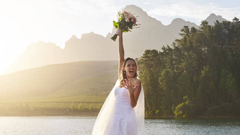 Eine Braut steht bei ihrer Hochzeit auf einem Steg an einem See und hält ihren Brautstrauß in die Höhe. Sie freut sich, weil sie bei ihrer Hochzeit mit günstigen DIY-Tipps Geld sparen konnte.