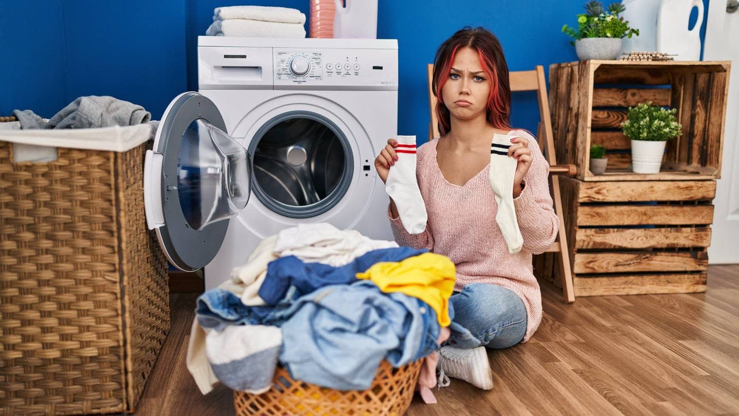 Eine Frau sitz mit einem Wäschekorb vor ihrer Waschmaschine, hält zwei unterschiedliche Socken in den Händen und macht ein trauriges Gesicht. Ist es wirklich so, dass Waschmaschinen Socken fressen? Wir klären im Faktencheck, warum sie manchmal darin verschwinden. (Foto: Adobe Stock, Krakenimages.com)
