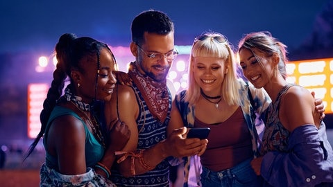 Gruppe von jungen Leuten ist zusammen auf einem Festival, im Hintergrund eine Bühne. Sie schauen gemeinsam auf ein Smartphone. Symbolbild für Musik-News und Ticket-Verlosungen im Whatsapp-Kanal von SWR3.