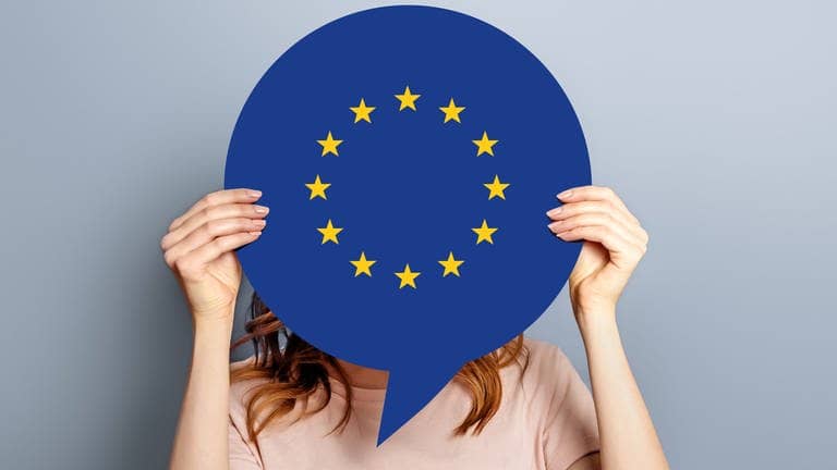 Eine Frau hält eine Spprechbalse vor ihrem Gesicht hoch, auf der das Zeichen für das Europäische Parlament zu sehen ist und fragt sich, was es mit ihr zu tun hat