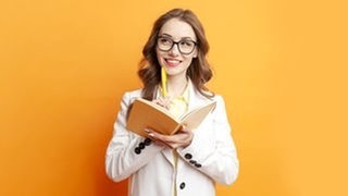Junge Frau hält denkend einen Stift ans Kinn und einen Schreib-Block in der Hand. Symbolbild für einen Artikel zum Thema Komma und Sätze, deren Bedeutung sich verändert, wenn man die Kommata verschiebt.