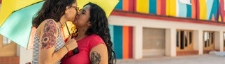 Zwei Personen stehen unter einem bunten Schirm und geben sich einen Paarbestätigungskuss. 