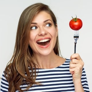 Junge Frau schaut erstaunt auf eine Tomate, die sie auf die Gabel gespießt hat. Symbolbild für den Unterschied zwischen Obst und Gemüse.