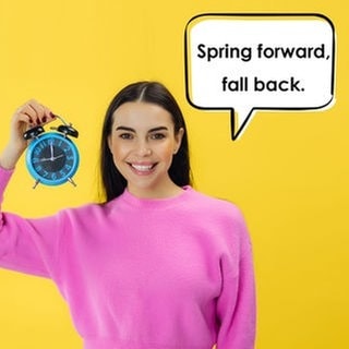 Eine junge Frau hält einen Wecker hoch und lächelt. Der Wecker zeigt 9:00 Uhr als Uhrzeit an. In einer Sprechblase neben ihr steht: „Spring forward, fall back.“ Symbolbild die Zeitumstellung