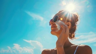Eine Frau schmiert sich Sonnencreme ins Gesicht. Im Hintergrund blauer Himmel und Sonne. Symbolfoto für den Test von Sonnencremes von Stiftung Warentest.
