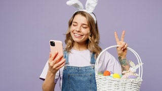 Junge Frau mit Smartphone in der Hand und Hasenohren auf dem Kopf hält einen Korb mit Ostereiern im Arm. Symbolbild für Lustige Whatsapp-Sprüche und Grüße zu Ostern.