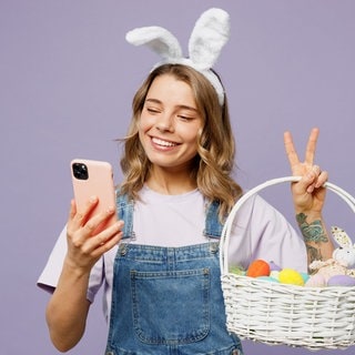 Junge Frau mit Smartphone in der Hand und Hasenohren auf dem Kopf hält einen Korb mit Ostereiern im Arm. Symbolbild für Lustige Whatsapp-Sprüche und Grüße zu Ostern.