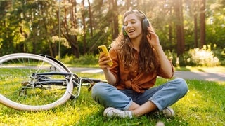 Junge Frau mit Kopfhörern und einem Smartphone in der Hand hört Musik. Sie sitzt im Gras, neben sich ein Fahrrad.