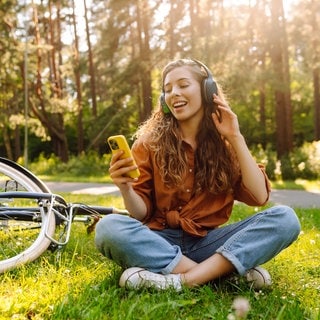 Junge Frau mit Kopfhörern und einem Smartphone in der Hand hört Musik. Sie sitzt im Gras, neben sich ein Fahrrad.