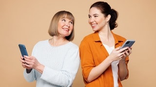 Mutter und Tochter mit einem Smartphone in der Hand, lächeln sich an. Symbolbild für lustige und schöne Whatsapp-Sprüche und Grüße an Mama zum Muttertag.