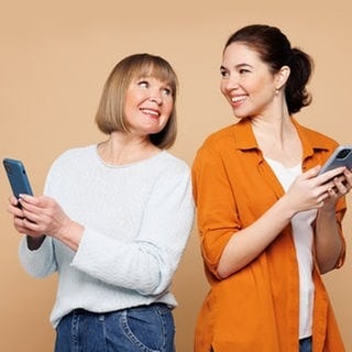 Mutter und Tochter mit einem Smartphone in der Hand, lächeln sich an. Symbolbild für lustige und schöne Whatsapp-Sprüche und Grüße an Mama zum Muttertag.