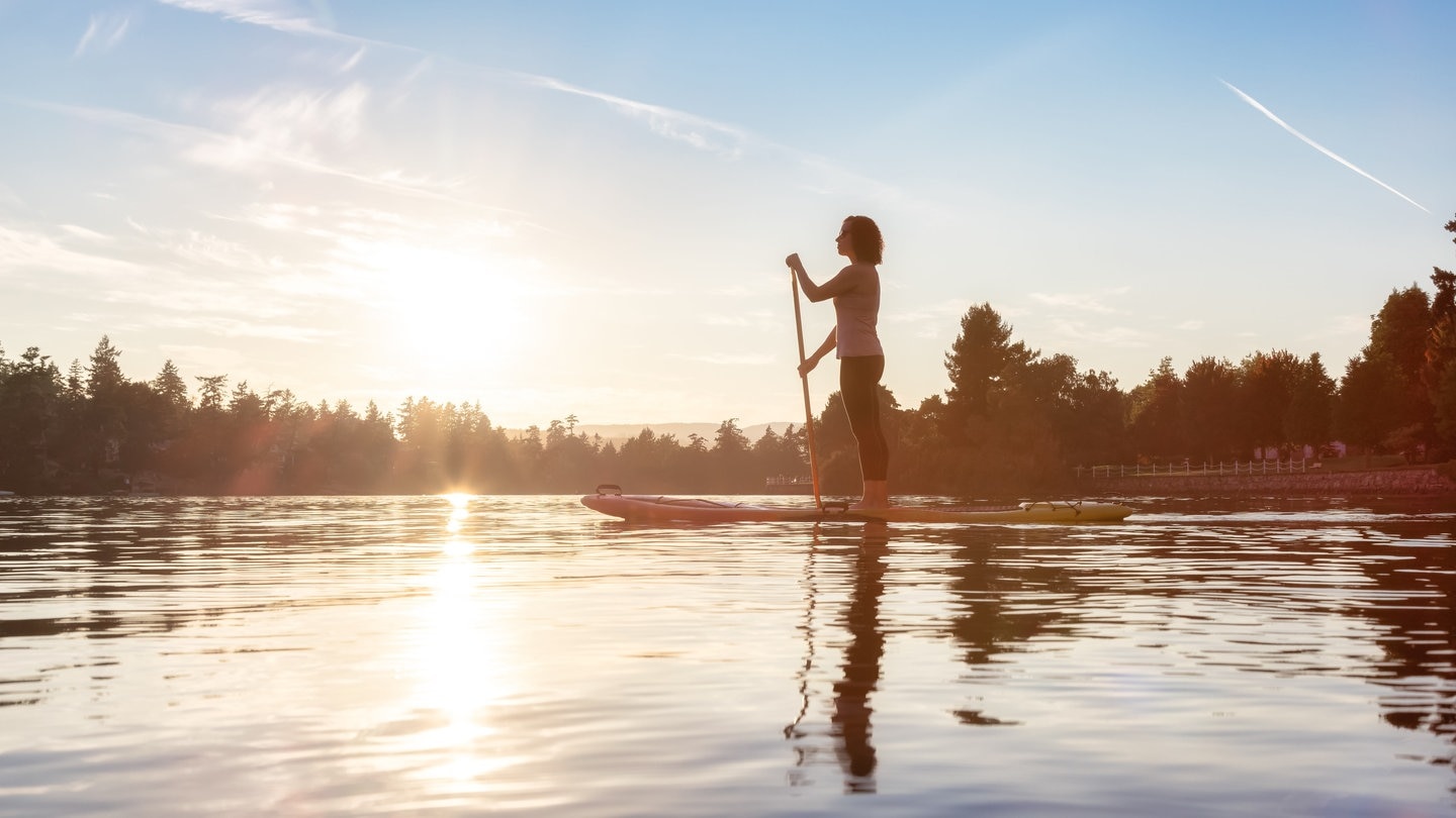 Eine Frau steht auf einem Stand-Up-Paddle-Board und paddelt auf einem See vor einer Waldlandschaft im Sonnenaufgang. Stiftung Warentest hat 10 SUP-Boards getestet.