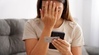 Eine junge Frau hält eine Hand vors Gesicht, in der anderen Handy und Kreditkarte. Sie ist auf einen Betrug reingefallen.