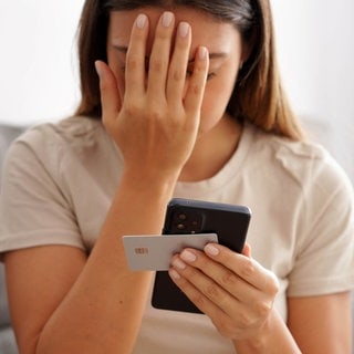 Eine junge Frau hält eine Hand vors Gesicht, in der anderen Handy und Kreditkarte. Sie ist auf einen Betrug reingefallen.