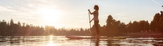 Eine Frau steht auf einem Stand-Up-Paddle-Board und paddelt auf einem See vor einer Waldlandschaft im Sonnenaufgang. Stiftung Warentest hat 10 SUP-Boards getestet. 