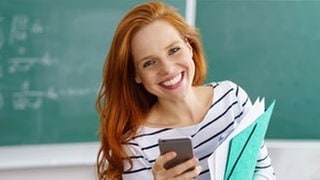 Lehrerin lacht vor einer Tafel in der Schule, in der Hand hält sie ein Smartphone und Hefte. Symbolbild für lustige Sprüche von Lehrern.