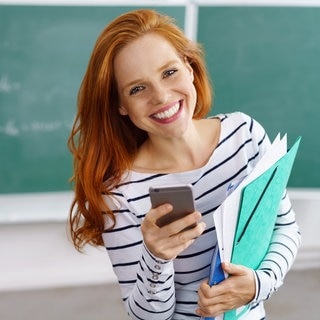 Lehrerin lacht vor einer Tafel in der Schule, in der Hand hält sie ein Smartphone und Hefte. Symbolbild für lustige Sprüche von Lehrern.