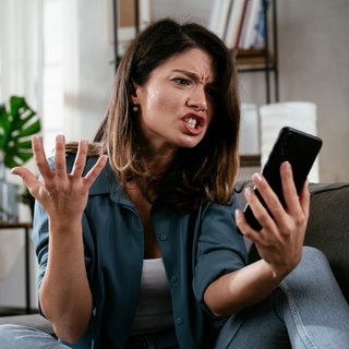 Eine Frau schaut wütend auf ihr Telefon. Betrugsanrufe und Spamanrufe – so könnt ihr euch vor Betrug schützen!