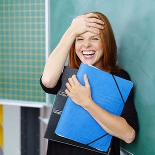 Lehrerin lacht vor einer Tafel in der Schule, in der Hand hält sie Hefte. Symbolbild für lustige Sprüche von Lehrern.