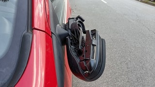 Ein Außenspiegel hängt beschädigt an einem roten Auto herunter. Betrüger zocken Autofahrer teilweise mit präparierten Außenspiegeln ab.