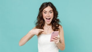 Junge Frau im Brautkleid zeigt lachend auf ihr Smartphone. Symbolbild für lustige Whatsapp-Sprüche und Glückwünsche zur Hochzeit.