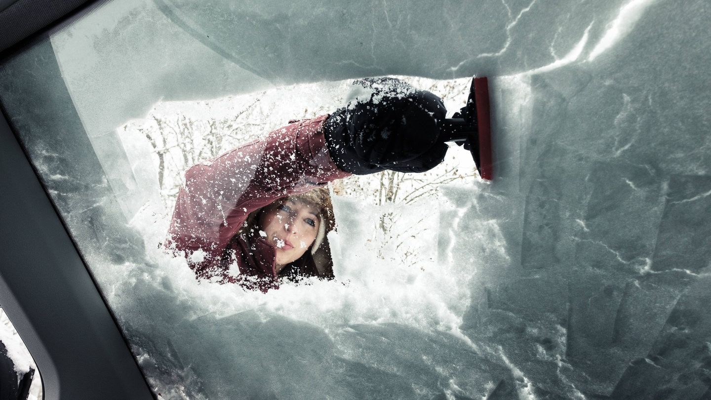 Junge Frau mit Enteiser, sie kratzt die verschneite und gefrorene Scheibe des Autos im Winter frei. (Foto: IMAGO, IMAGO / Westend61)