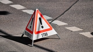 Eine mobile, dreieckige Warnpyramide steht auf einer asphaltierten Straße mit Markierungen. Sie verweist auf Stau.