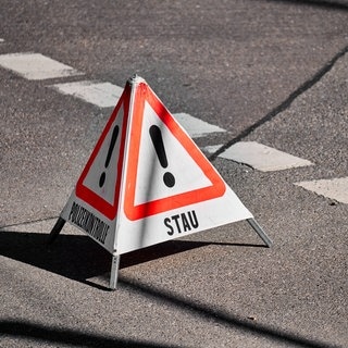 Eine mobile, dreieckige Warnpyramide steht auf einer asphaltierten Straße mit Markierungen. Sie verweist auf Stau.