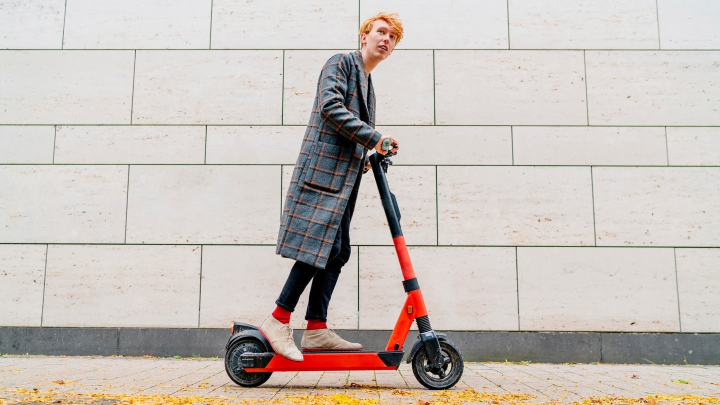 Mann mit E-Scooter auf Bürgersteig vor grauer Wand (Foto: dpa Bildfunk, Picture Alliance)