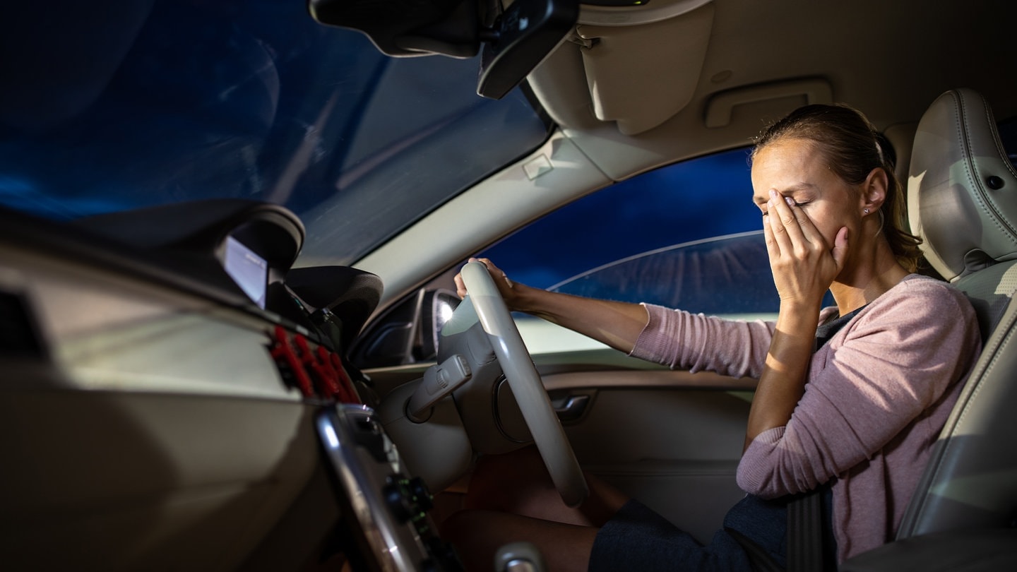 Eine Frau sitzt am Steuer und verfällt in einen Sekundenschlaf, was gefährlich enden kann (Foto: Adobe Stock, lightpoet)