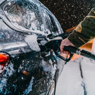 Ein schwarzes Auto wird im Winter an einer Tankstelle betankt. Es fallen Schneeflocken. Das Foto soll den erhöhten Spritverbrauch im Winter, beispielsweise auf dem Weg in den Skiurlaub, verdeutlichen. Wir klären, woran das liegt und welche Tipps es gibt, um die größtens Spritfresser im Winter zu vermeiden.
