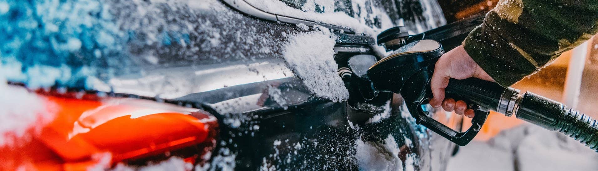 Ein schwarzes Auto wird im Winter an einer Tankstelle betankt. Es fallen Schneeflocken. Das Foto soll den erhöhten Spritverbrauch im Winter, beispielsweise auf dem Weg in den Skiurlaub, verdeutlichen. Wir klären, woran das liegt und welche Tipps es gibt, um die größtens Spritfresser im Winter zu vermeiden.
