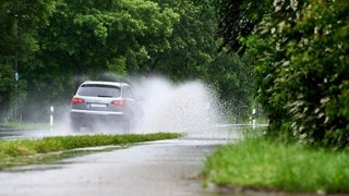 Ein silbernes Auto fährt vorsichtig bei Nässe über eine überflutete Straße, denn es besteht Gefahr durch Aquaplaning