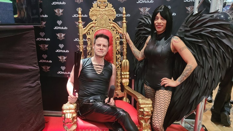 Voller Körpereinsatz: Unser Reporter Christian Karsch auf der „Obscene“-Erotik-und-Fetisch-Messe in Karlsruhe. Neben ihm eine Frau in schwarzem Lederkostüm und mit schwarzen Englesflügeln. (Foto: SWR)