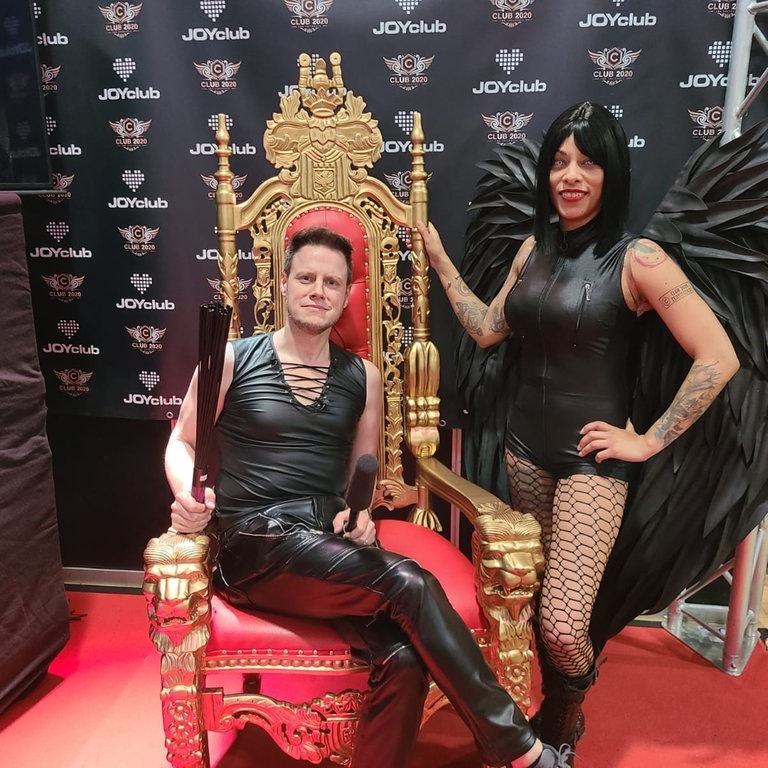 Voller Körpereinsatz: Unser Reporter Christian Karsch auf der „Obscene“-Erotik-und-Fetisch-Messe in Karlsruhe. Neben ihm eine Frau in schwarzem Lederkostüm und mit schwarzen Englesflügeln. (Foto: SWR)