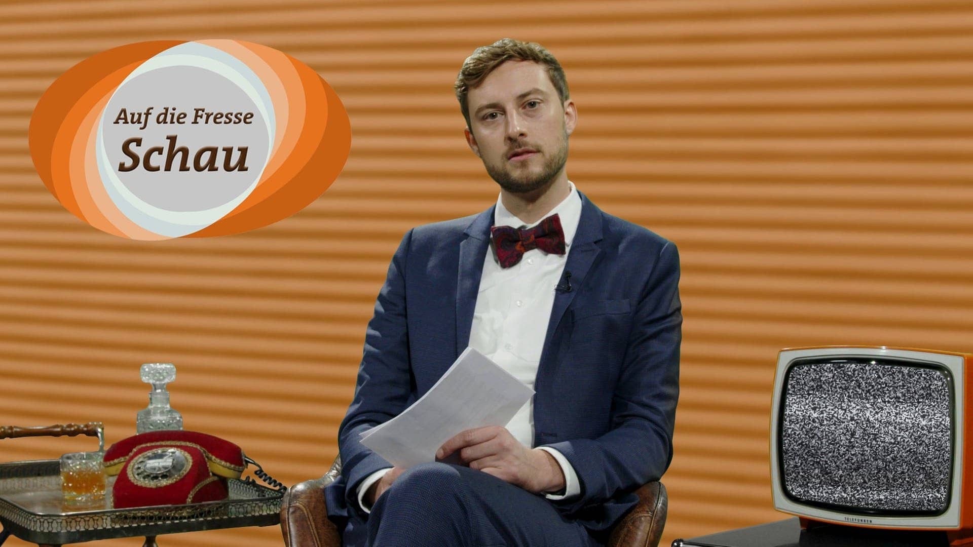 SWR3-Moderator Constantin Zöller sitzt mit Jackett in einem Fernsehstudio. Er liest Hasskommentare, das Logo der Sendung ist eingeblendet mit der Aufschrift „Auf-die-Fresse“-Schau. (Foto: SWR3)