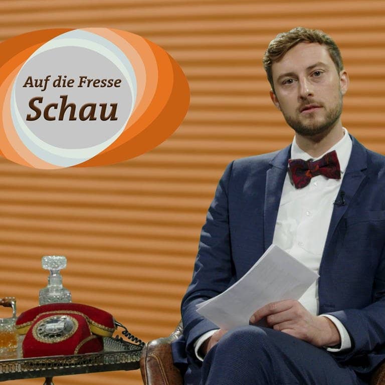 SWR3-Moderator Constantin Zöller sitzt mit Jackett in einem Fernsehstudio. Er liest Hasskommentare, das Logo der Sendung ist eingeblendet mit der Aufschrift „Auf-die-Fresse“-Schau. (Foto: SWR3)