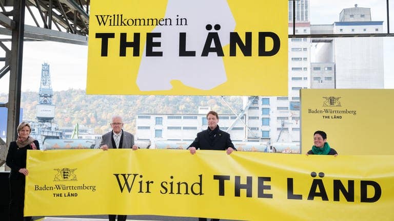 Poltiker halten Banner mit Baden-Württembergs Slogan "The Länd" hoch.