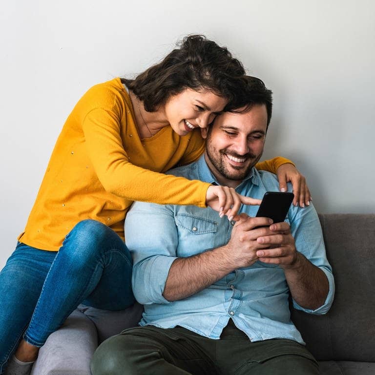 Junge Frau und junger Mann sitzen lachend auf der Couch. Sie zeigt auf der Smartphone, sie gucken sich vermutlich die Top10 der lustigsten Videos des Jahres von SWR3 an. (Foto: Adobe Stock, Tijana)