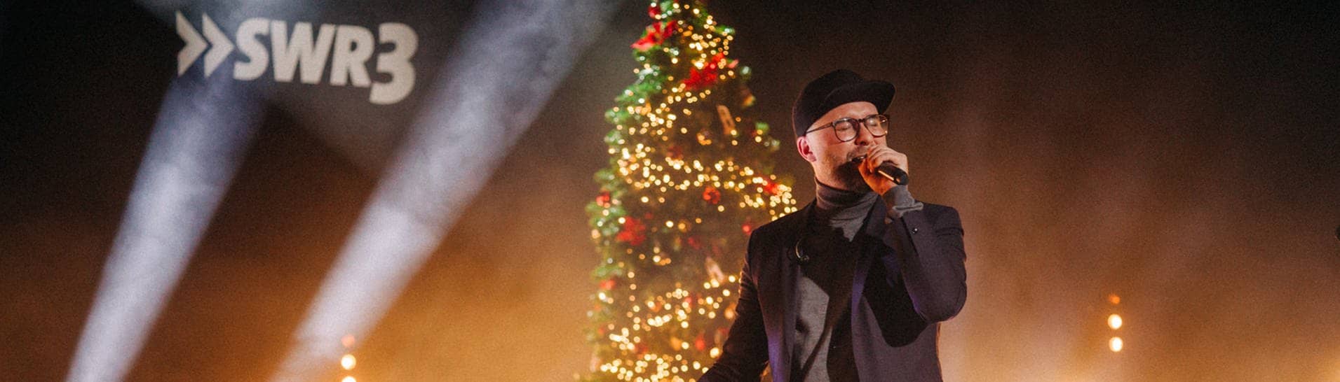 Mark Forster mit Cap und Mikro auf der Bühne vor einem beleuchteten Weihnachtsbaum (Foto: SWR3, SWR3/Niko Neithardt)