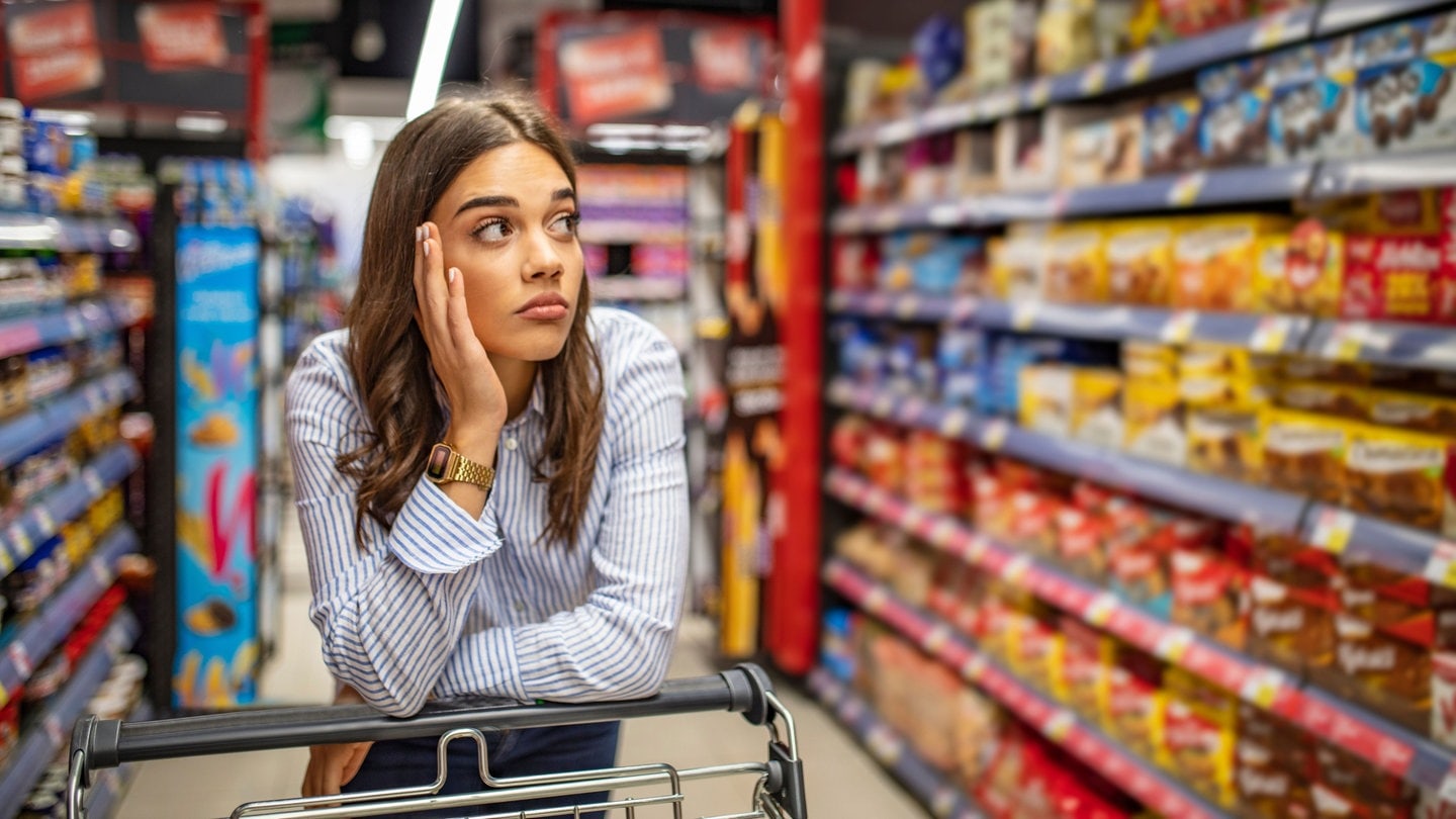 Eine Frau steht mit ihrem Wagen im Supermarkt und schaut frustriert die Regale an (Foto: Adobe Stock Fotos/Dragana Gordic)