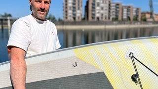 Christoph Rombach von LAKS Stand up paddling im Zollhafen Mainz hält eines der SUP-Board in der Hand, die er verleiht. Dahinter ist der Neckar in Mannheim und Hochhäuser zu sehen. Stiftung Warentest hat getestet, welche Stand-Up-Paddle richtig gut sind (Foto: SWR, Jakob Reifenberger) Christoph Rombach von LAKS Stand up paddling im Zollhafen Mainz hält eines der SUP-Board in der Hand, die er verleiht. Dahinter ist der Neckar in Mannheim und Hochhäuser zu sehen. Stiftung Warentest hat getestet, welche Stand-Up-Paddle richtig gut sind