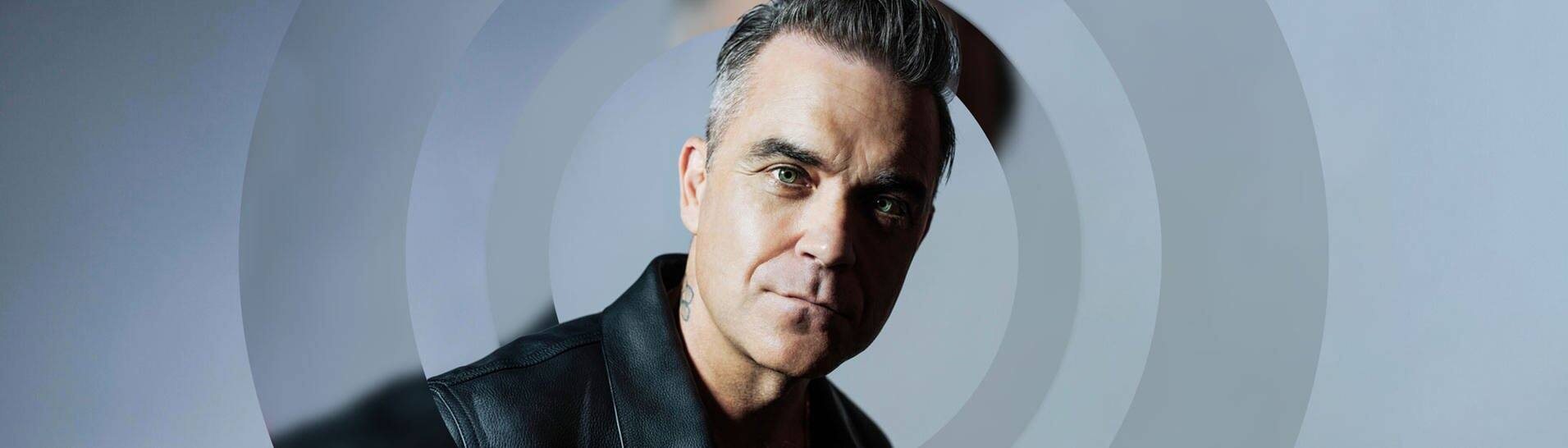 Der Sänger Robbie Williams vor einem blauen Hintergrund, er trägt eine schwarze Lederjacke. (Foto: Leo Baron)
