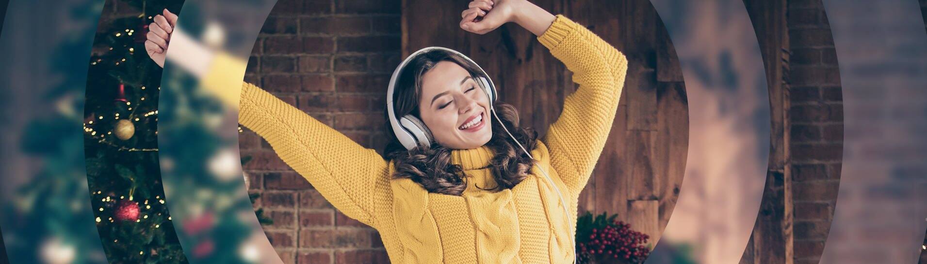 Eine junge Frau in einem gelben Pullover tanzt vor einem Weihnachtsbaum und hört Musik mit Kopfhörern. (Foto: Adobe Stock / deagreez)
