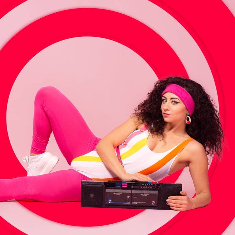Eine Frau mit schwarzen Haaren und einem rosa Stirnband liegt in einem Sport.Outfit auf dem Boden und hält ihre Hand auf einem Kasettenrecorder (Foto: Adobe Stock / yuliiapedchenko)