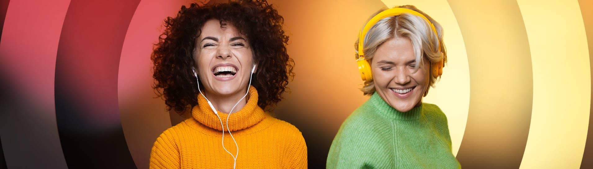 Eine schwarzhaarige und und blonde Frau hören mit Kopfhörern Musik, tanzen und lachen.