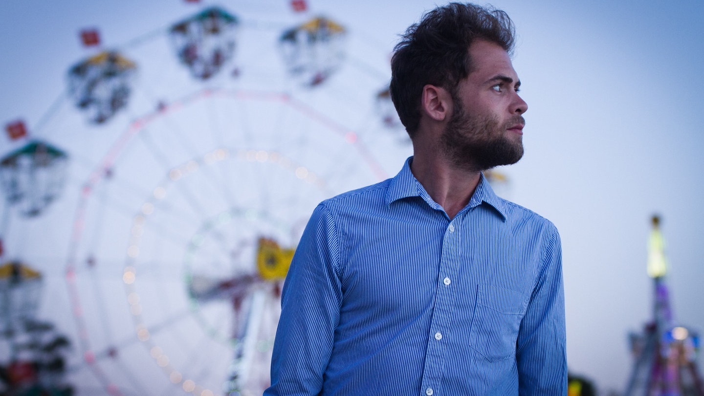 Der Singer-Songwriter Passenger trägt ein weiß-blau gestreiftes Hemd und steht vor einem Riesenrad, sein größter Hit ist „Let Her Go“ (Foto: Angelo Kehagias)