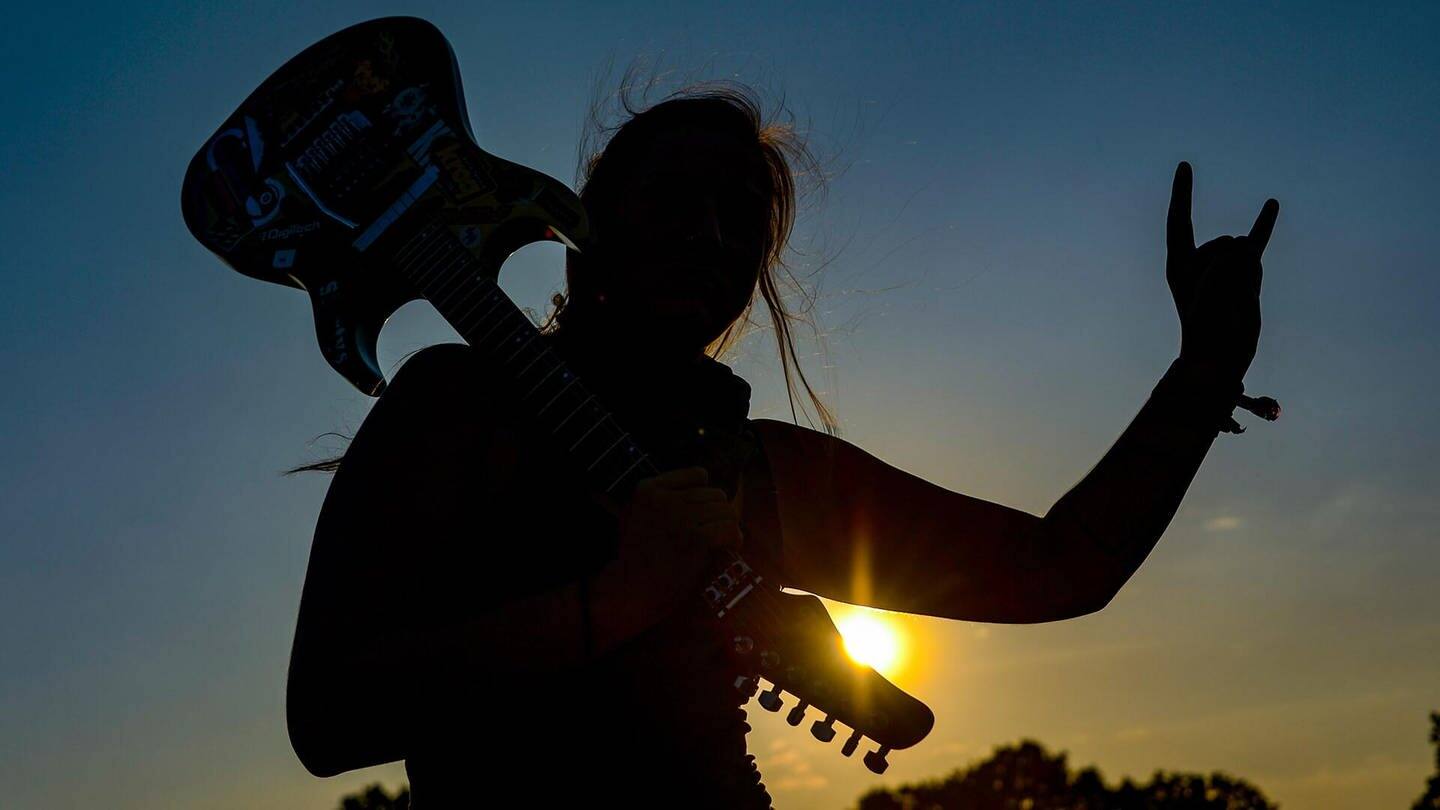 Frau mit E-Gitarre auf der Schulter zeigt den Metalgruß, die Pommesgabel. (Foto: picture alliance/Axel Heimken/dpa)
