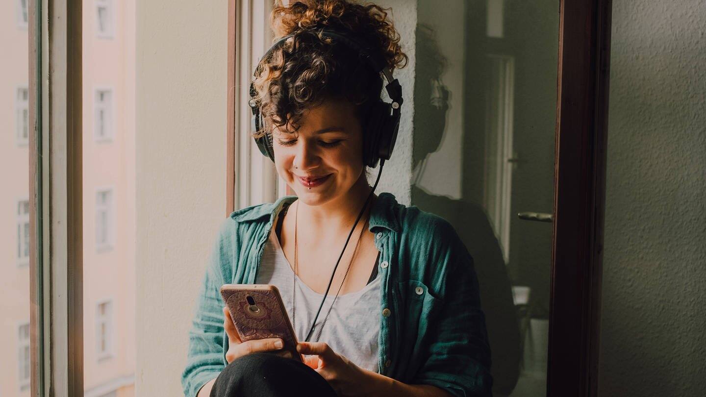 Junge Frau sitzt am Fenster, hat Kopfhörer auf und lächelt ihr Handy an (Foto: imago images, Addictive Stock)